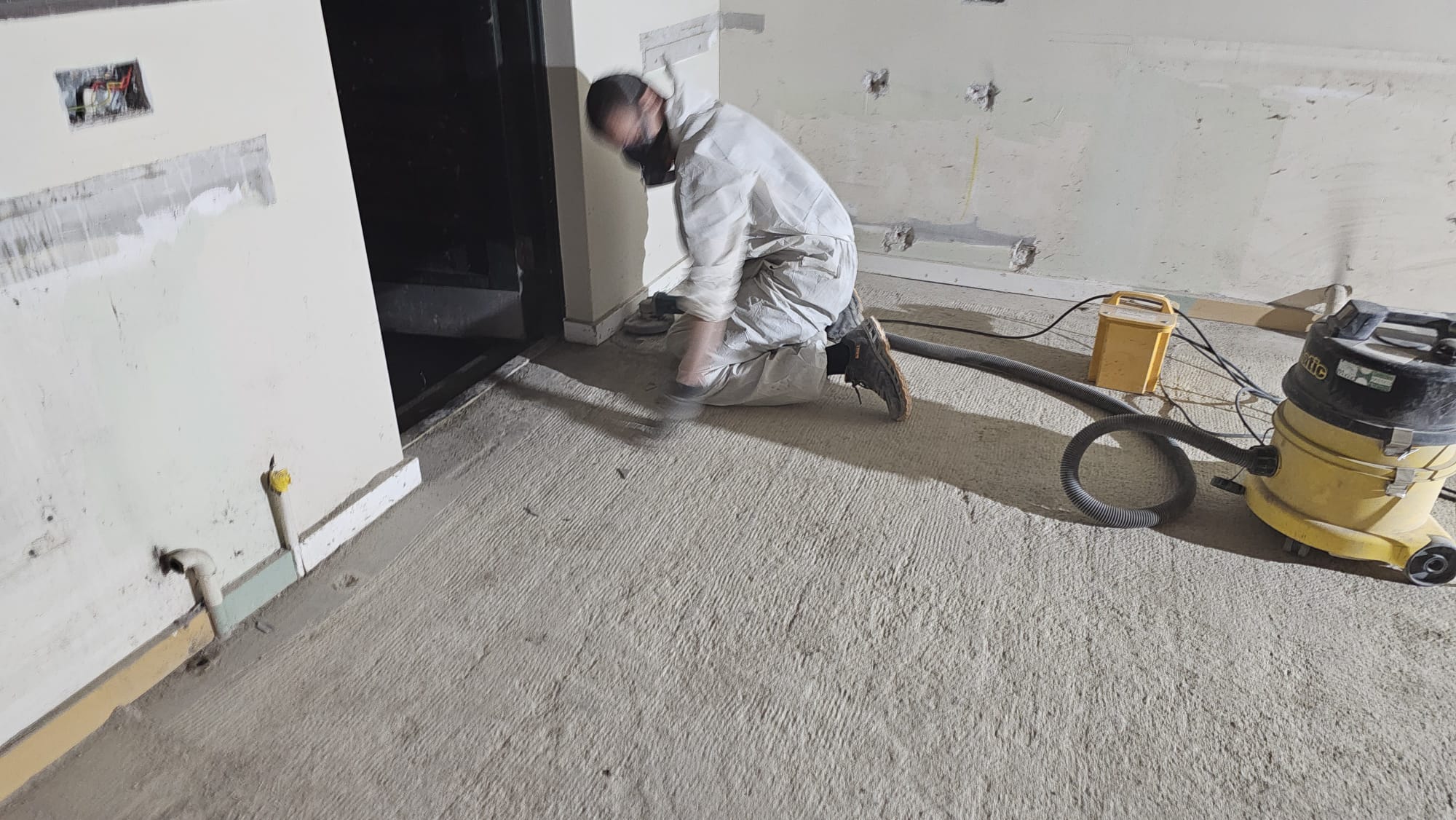 Worker in white protective suit using a floor grinder with dust extraction during controlled asbestos work